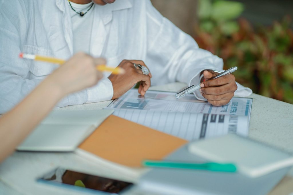 Crop anonymous multiracial female friends sitting at table together while studying with books and pen in daytime