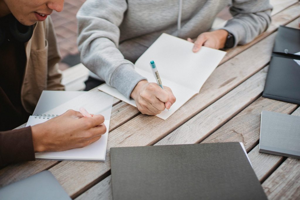 High angle crop male students in casual clothes taking notes in copybooks while working on home assignment together in park